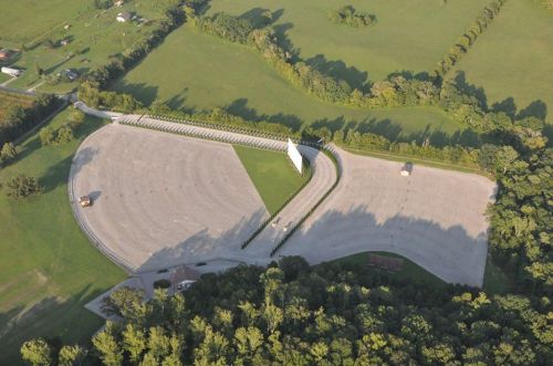 Aerial view of a large, empty parking lot surrounded by green fields and trees, with a white structure in the center.