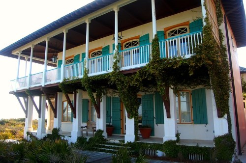 A coastal house with green shutters, white columns, and climbing vines, surrounded by lush vegetation.