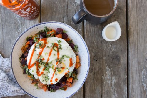 A bowl of sautéed vegetables topped with a fried egg and sriracha, alongside a cup of coffee and a small creamer.