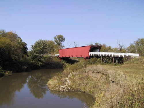 A red covered bridge spans a calm river, surrounded by trees and grassy banks under a clear blue sky.