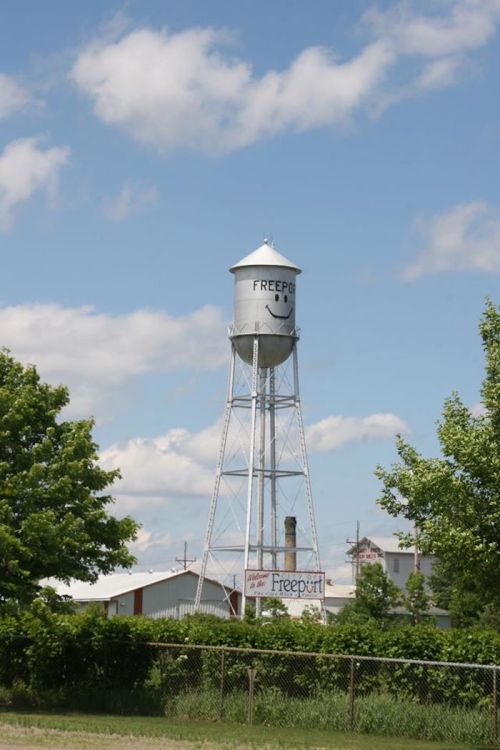 A silver water tower with "FREEPORT" written on it, surrounded by green trees and a blue sky with clouds.