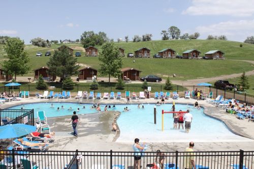 A sunny pool area with people swimming and lounging, surrounded by cabins and green hills in the background.