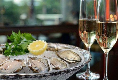 A platter of fresh oysters on ice, garnished with lemon and parsley, alongside two glasses of sparkling wine.