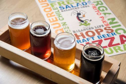A wooden tray holds four glasses of different craft beers, with a colorful menu in the background.