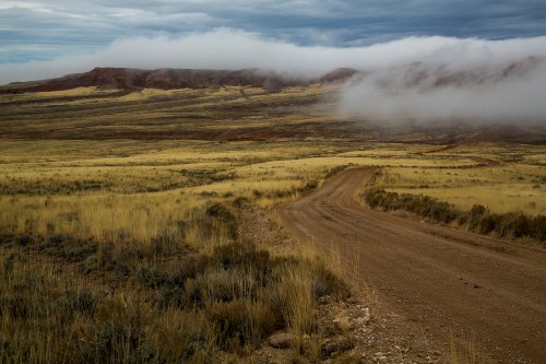 A winding dirt road leads through a grassy landscape with low clouds and distant hills under a cloudy sky.