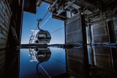 A ski gondola reflected in a puddle, framed by a mountain station under a clear blue sky.