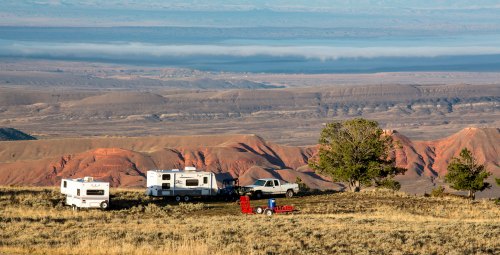 A scenic view of RVs parked on a hillside, overlooking colorful canyons and distant mountains under a clear sky.