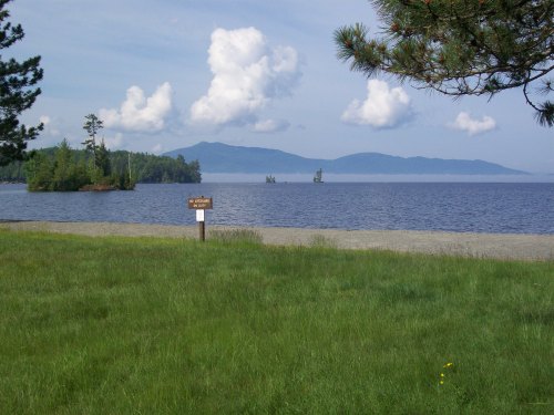 A serene lake view with grassy shore, distant mountains, and fluffy clouds in a clear blue sky.