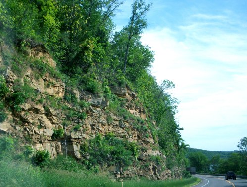 A winding road alongside a rocky cliff covered in lush greenery under a clear blue sky.