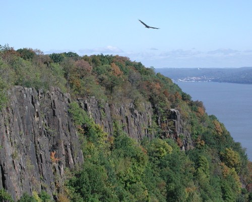 A scenic view of a rocky cliffside with colorful trees and a river in the background, under a clear blue sky.