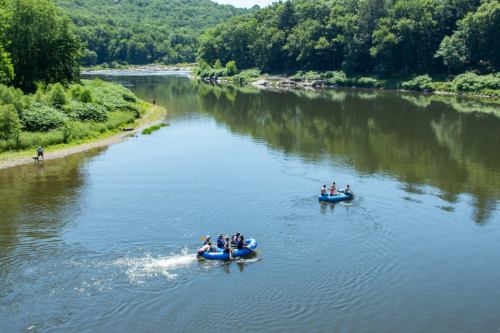 Two groups of people in blue rafts float on a calm river surrounded by lush green trees and hills.