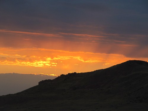 A dramatic sunset casts orange and yellow hues over a silhouetted hillside against a cloudy sky.