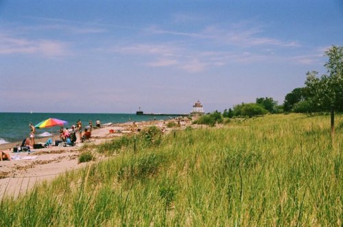 A sunny beach scene with people sunbathing, colorful umbrellas, and a lighthouse in the distance along the shoreline.