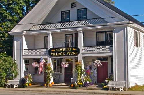 A charming white building with a sign reading "Newbury Village Store," adorned with flowers and pumpkins.