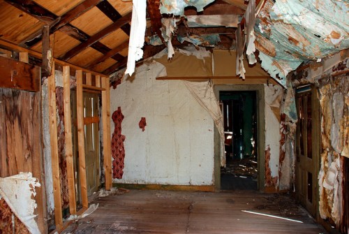 Interior of a dilapidated room with peeling walls, exposed wood, and a damaged ceiling, showcasing signs of neglect.