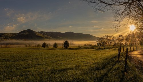 A serene landscape at sunrise, featuring misty mountains, green fields, and a tree-lined road.