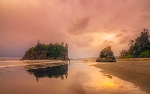 A serene beach scene at sunset, featuring rocky formations and reflections in the wet sand under a colorful sky.