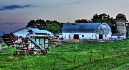 A rustic farm scene featuring a vintage combine harvester, a barn, and a silo surrounded by green fields and trees.