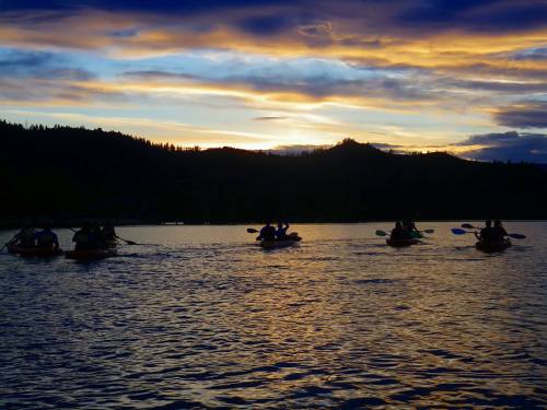 Silhouettes of kayakers on a lake at sunset, with vibrant clouds and mountains in the background.