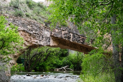 A natural stone arch spans over a river, surrounded by lush greenery and rocky terrain.