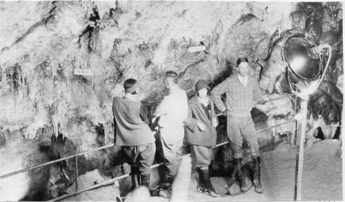 Four people exploring a cave, surrounded by stalactites and stalagmites, with a large lamp illuminating the scene.