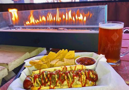 A hot dog topped with condiments, served with tortilla chips and salsa, alongside a beer and a flame feature in the background.