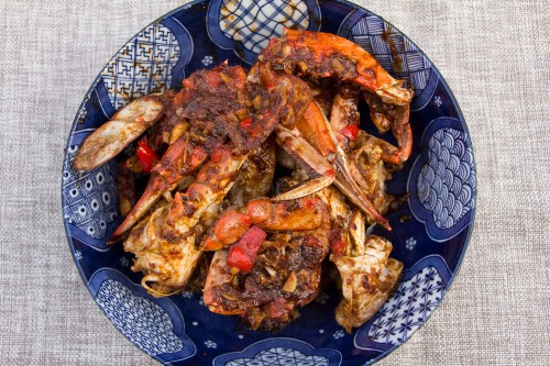 A blue bowl filled with spicy, cooked crabs garnished with red peppers and herbs on a textured beige surface.