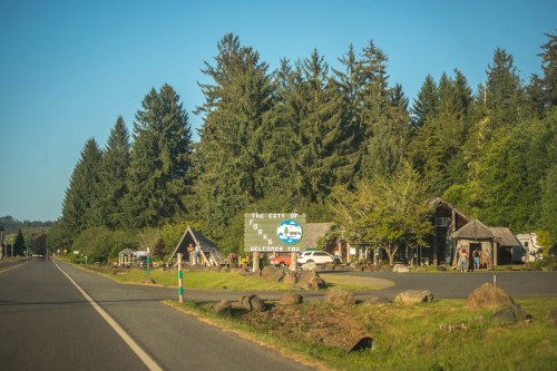 A welcome sign for the city of Forks, surrounded by trees and rustic buildings along a quiet road.