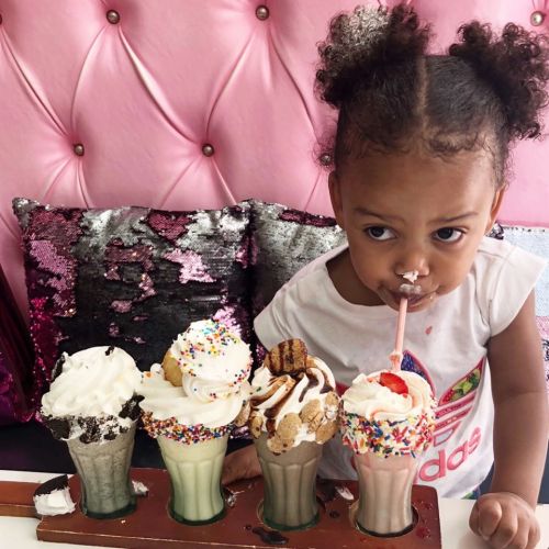A child with curly hair enjoys milkshakes topped with whipped cream and sprinkles, sitting in a colorful setting.
