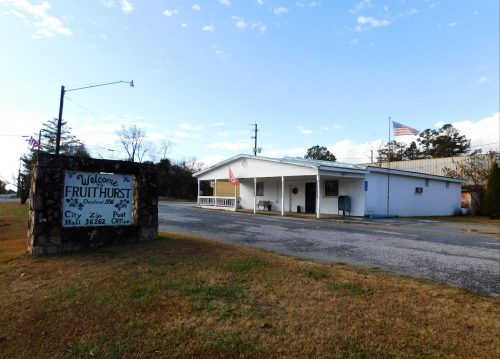 A small building with a sign reading "Welcome to Fruithurst" and an American flag, set against a clear sky.