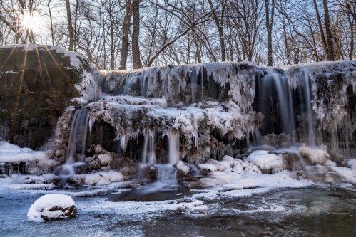 A frozen waterfall surrounded by icy rocks and trees, with sunlight shining through the branches above.