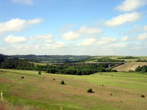 A scenic landscape featuring rolling green hills, a bridge in the distance, and a blue sky with fluffy clouds.