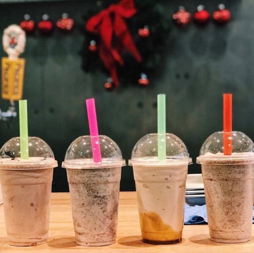 Four colorful milkshake cups with clear lids and straws, arranged on a wooden table, festive decorations in the background.