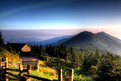 A scenic mountain landscape at dawn, featuring a wooden cabin, lush trees, and rolling hills under a colorful sky.