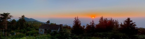 A panoramic view of a sunset over mountains, with a rustic house and trees in the foreground.
