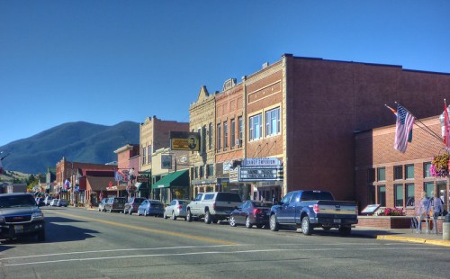 A charming small-town street lined with historic buildings, shops, and parked cars, set against a mountain backdrop.