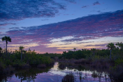 A serene landscape at dusk, featuring a calm waterway, lush greenery, and vibrant clouds in shades of purple and pink.