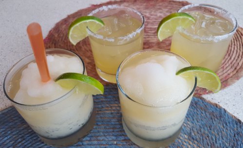Four refreshing cocktails with lime slices, one with a straw, served on a woven mat.
