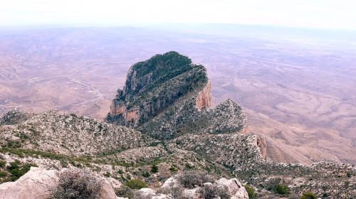 A rugged mountain peak rises above a vast, arid landscape under a cloudy sky.