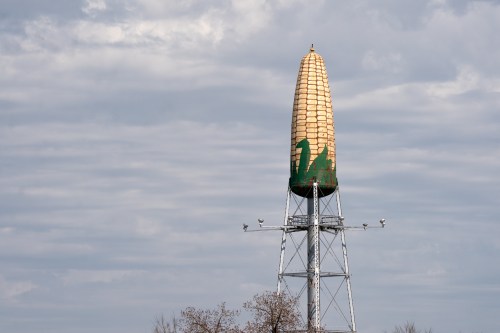 A tall water tower shaped like an ear of corn, painted green and gold, against a cloudy sky.