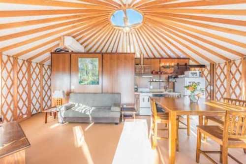 Interior of a cozy yurt with wooden beams, a sofa, dining table, and kitchen area, illuminated by natural light.