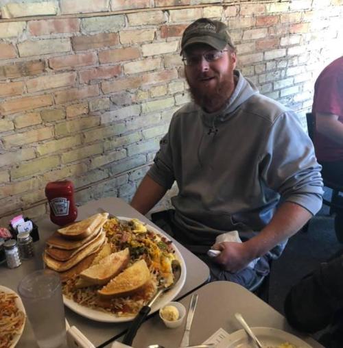 A man with a beard sits at a table with a large plate of tacos and a side of fries, smiling in a casual restaurant.