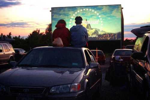 A couple sits on a car roof watching a movie at a drive-in theater as the sun sets in the background.