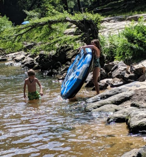 Two children play by a river; one is in the water while the other carries a blue inflatable tube.