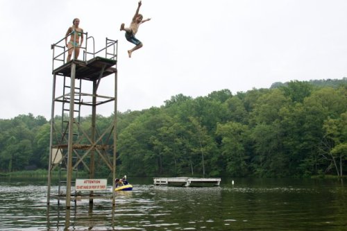 A boy jumps off a diving platform into a lake, while a girl watches from above and a boat floats nearby.