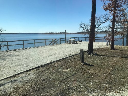 A serene lakeside view with a wooden deck, picnic table, and trees under a clear blue sky.