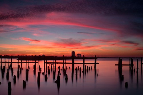 A serene sunset over water, with silhouetted pilings and colorful clouds reflecting in the calm surface.
