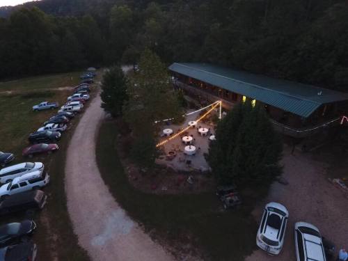 Aerial view of a rustic building with outdoor seating, surrounded by trees and parked cars along a winding gravel path.