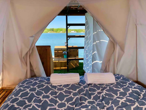 Cozy tent interior with a bed and towels, overlooking a serene lake view through an open entrance.