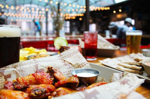 A table with chicken wings, dipping sauce, drinks, and snacks in a lively restaurant setting with string lights.